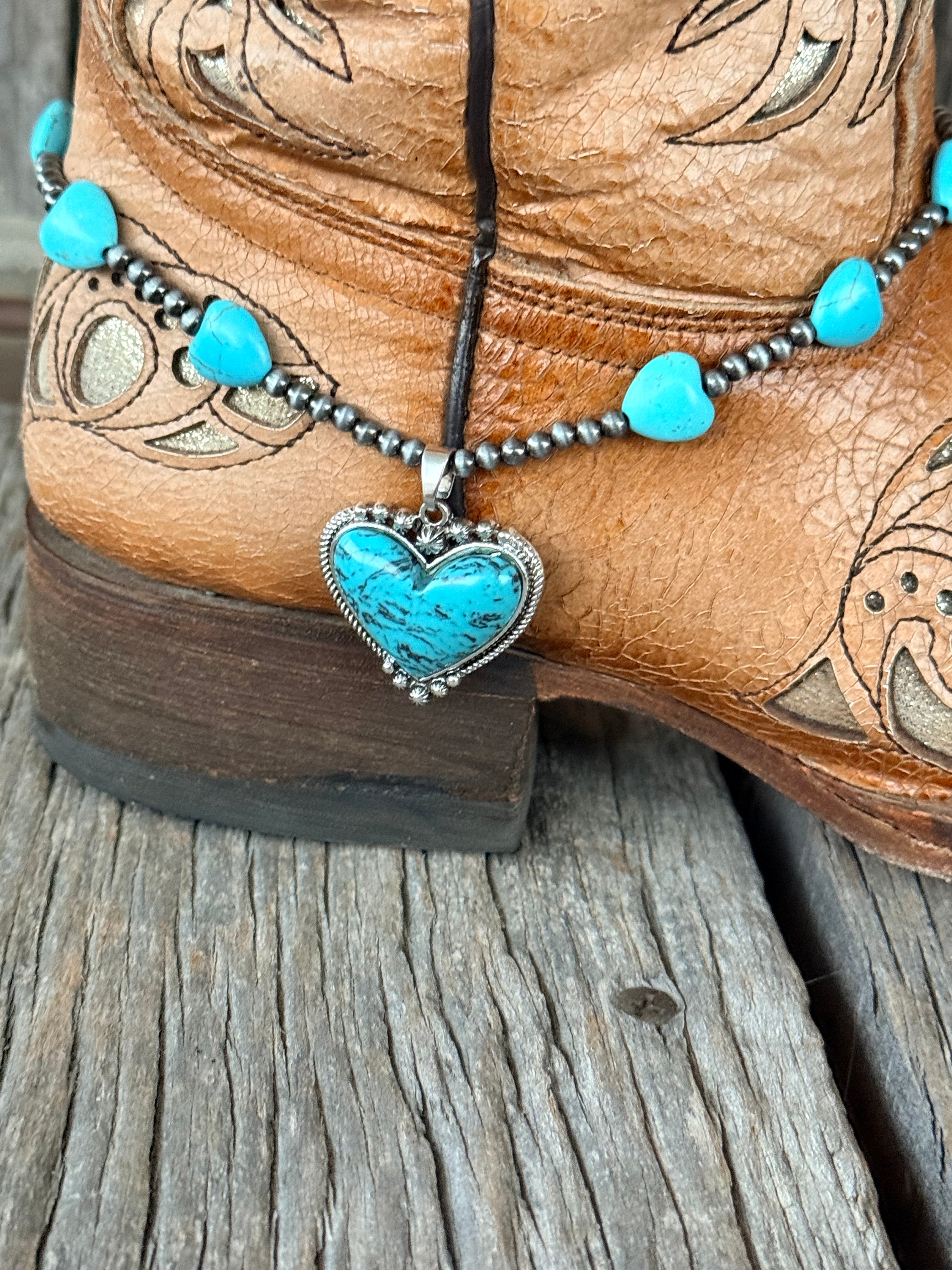 Front low angle view of turquoise heart bead boot bracelet with large turquoise heart stone pendant with rope-twist silver border and silver star accent draping at ankle of tan tooled cowboy boot
