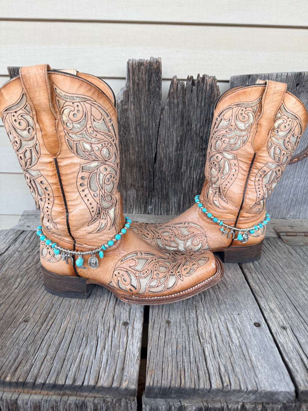 Turquoise bead and silver charm boot bracelet with Good Luck horseshoe and Western charm cluster worn on tan tooled laser-cut cowboy boots, full pair front view on wood surface
