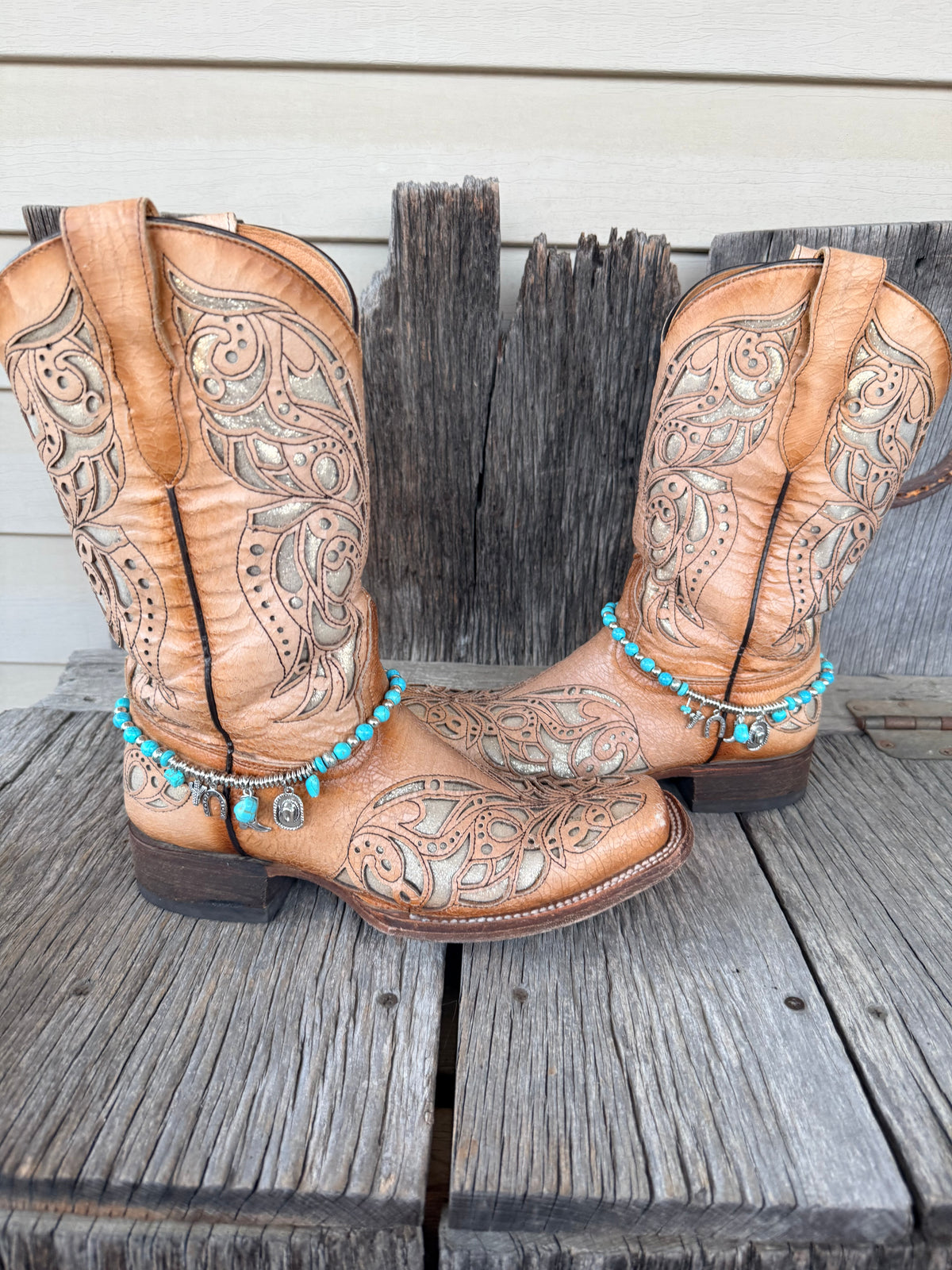 Turquoise bead and silver charm boot bracelet with Good Luck horseshoe and Western charm cluster worn on tan tooled laser-cut cowboy boots, full pair front view on wood surface
