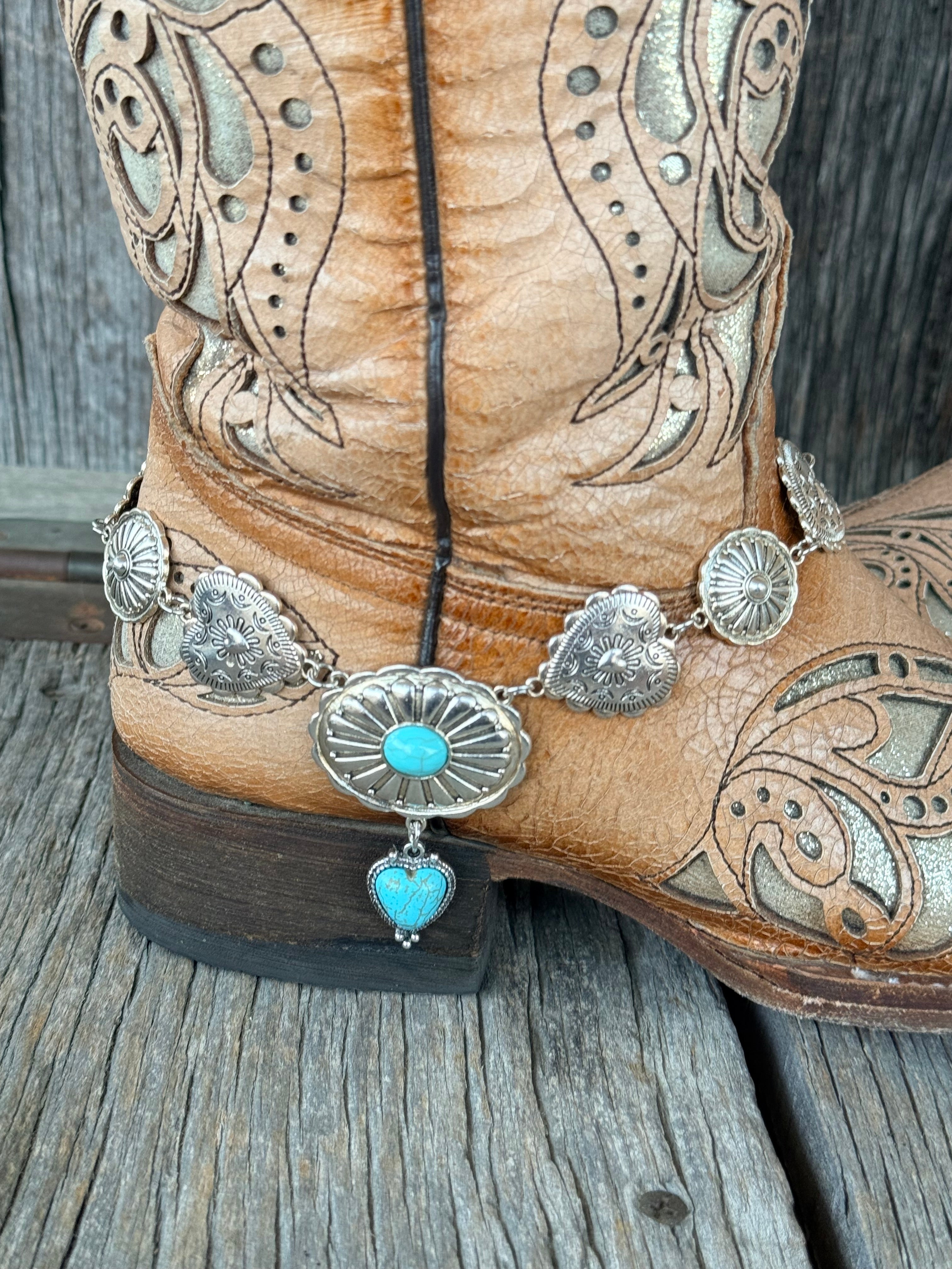 Close-up of linked silver concho chain boot bracelet with large oval sunburst concho centerpiece, turquoise cabochon stone, and turquoise heart charm drop at heel of tan tooled cowboy boot
