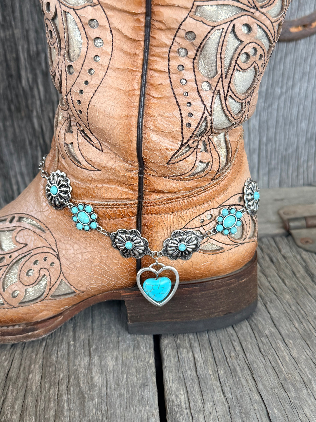 Side view of turquoise flower concho and oval turquoise cluster chain boot bracelet with open silver heart pendant drop at ankle of tan laser-cut tooled cowboy boot
