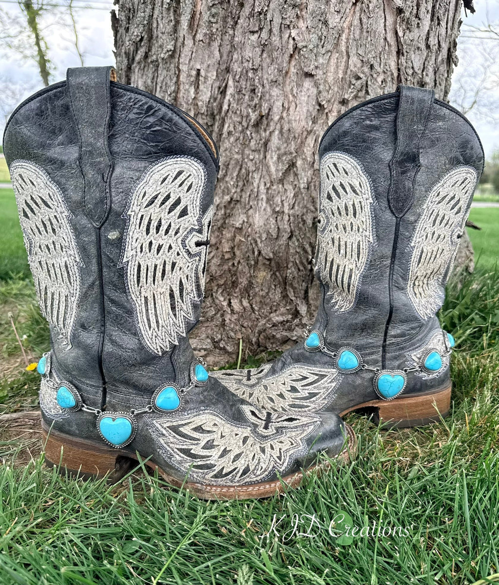 Turquoise heart boot bracelet with silver beaded bezel chain draped across the toe of black angel wing embroidered cowboy boots resting against a tree outdoors
