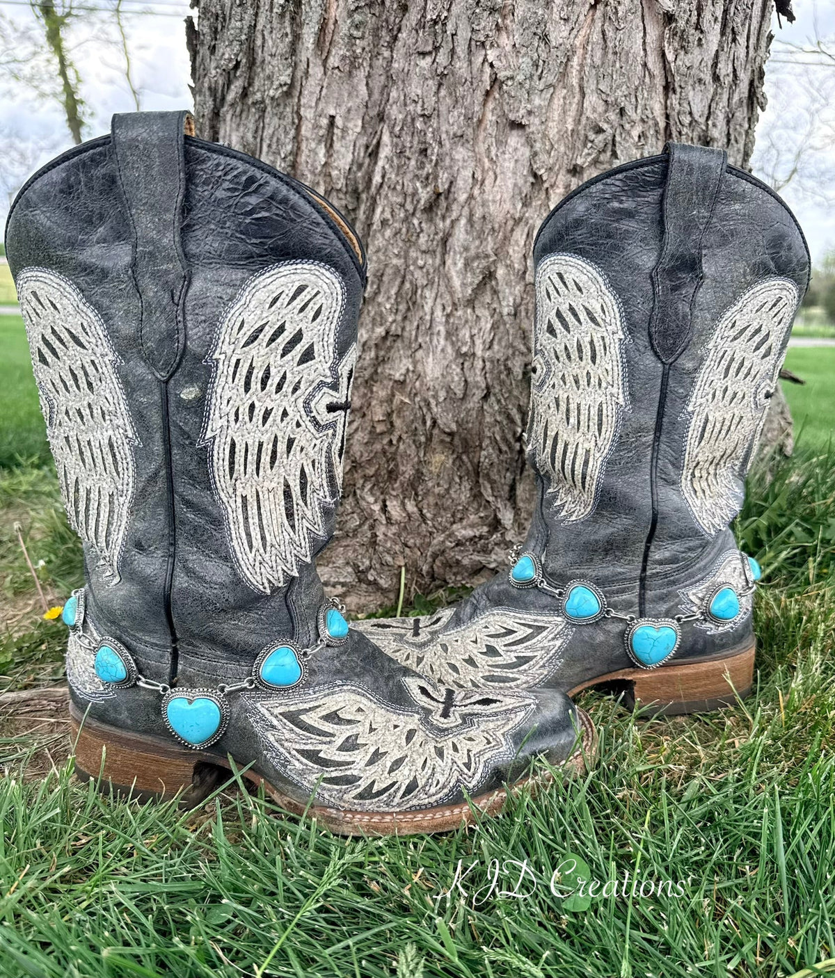 Turquoise heart boot bracelet with silver beaded bezel chain draped across the toe of black angel wing embroidered cowboy boots resting against a tree outdoors
