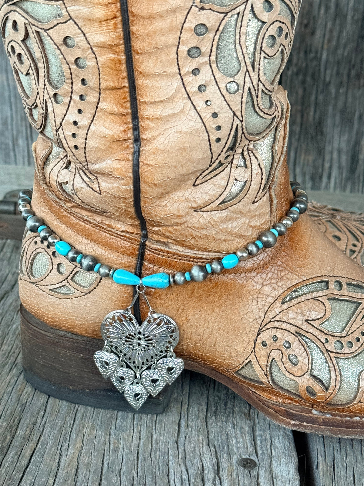 Side view of gunmetal Navajo pearl and turquoise teardrop boot bracelet with large silver filigree heart pendant draping at ankle of tan tooled laser-cut cowboy boot on wood surface
