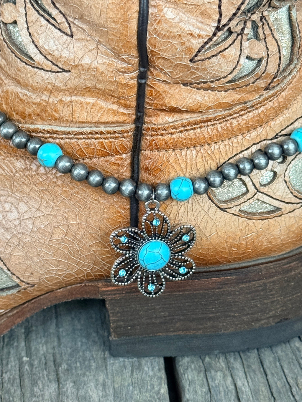 Detail view of large ornate silver flower concho pendant with turquoise cabochon center and turquoise crystal rhinestone petal accents on gunmetal Navajo pearl and turquoise bead boot bracelet

