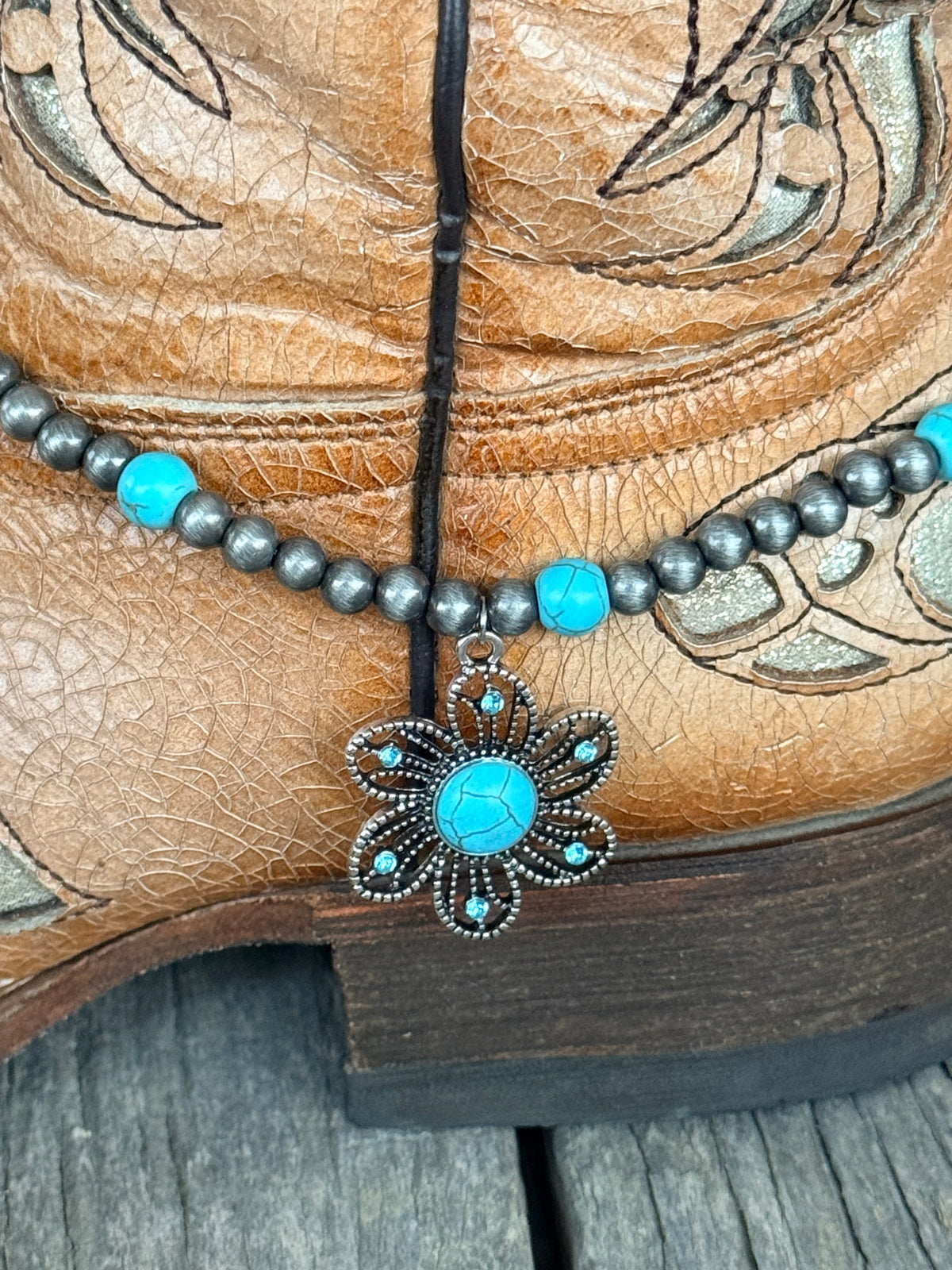 Detail view of large ornate silver flower concho pendant with turquoise cabochon center and turquoise crystal rhinestone petal accents on gunmetal Navajo pearl and turquoise bead boot bracelet
