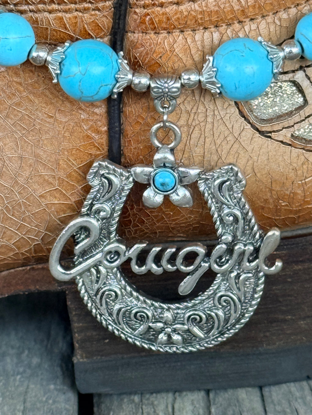 Detail view of large ornate silver horseshoe pendant with rope border, floral scroll engraving, Cowgirl script, and small silver flower charm with turquoise cabochon drop on turquoise bead boot bracelet
