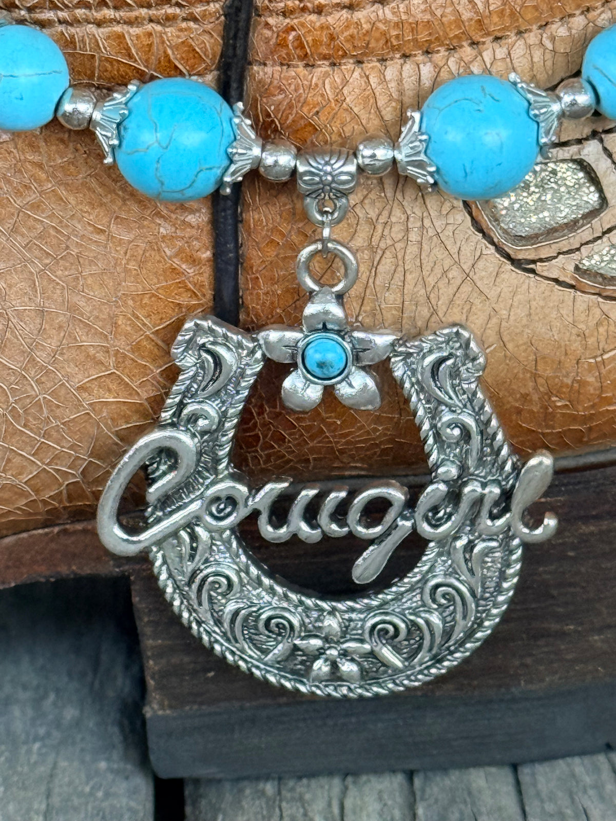 Detail view of large ornate silver horseshoe pendant with rope border, floral scroll engraving, Cowgirl script, and small silver flower charm with turquoise cabochon drop on turquoise bead boot bracelet
