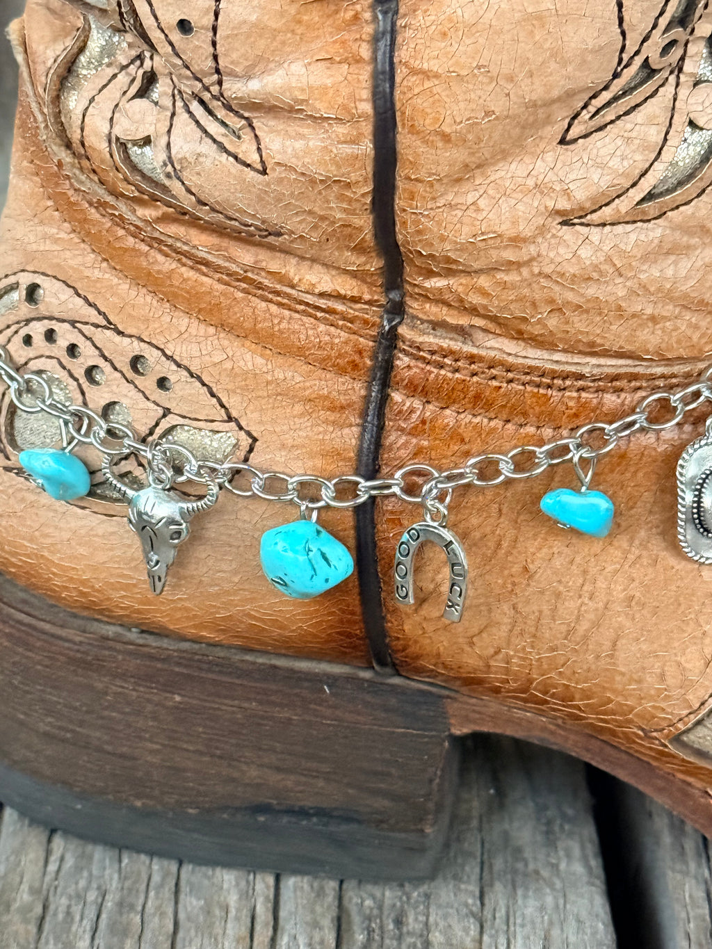 Detail close-up of silver Western charm boot bracelet showing turquoise stone chips, silver steer head charm, Good Luck horseshoe charm, and cowboy hat concho charm at heel of tan tooled cowboy boot
