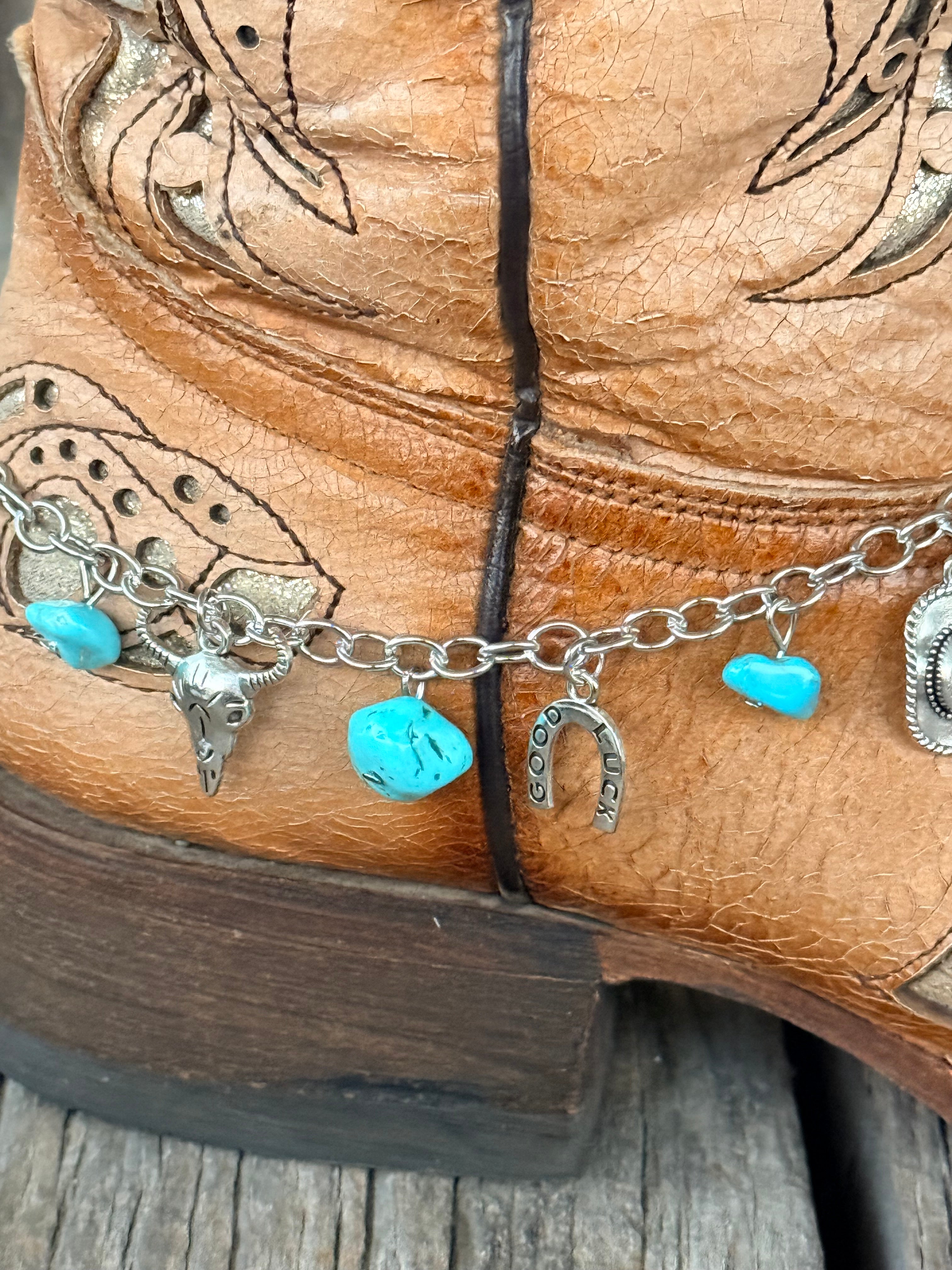 Detail close-up of silver Western charm boot bracelet showing turquoise stone chips, silver steer head charm, Good Luck horseshoe charm, and cowboy hat concho charm at heel of tan tooled cowboy boot
