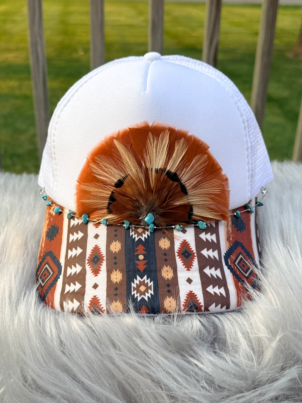 A white trucker-style cap with a Southwestern geometric patterned visor, decorated with a brown feather fan and turquoise bead accents,