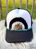 A black and white trucker hat with a decorative feather and turquoise bead embellishment on the brim,