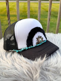 A black and white trucker hat with turquoise bead trim and a feathered decorative accent, resting on a white faux fur surface with a wooden fence and green grass in the background.