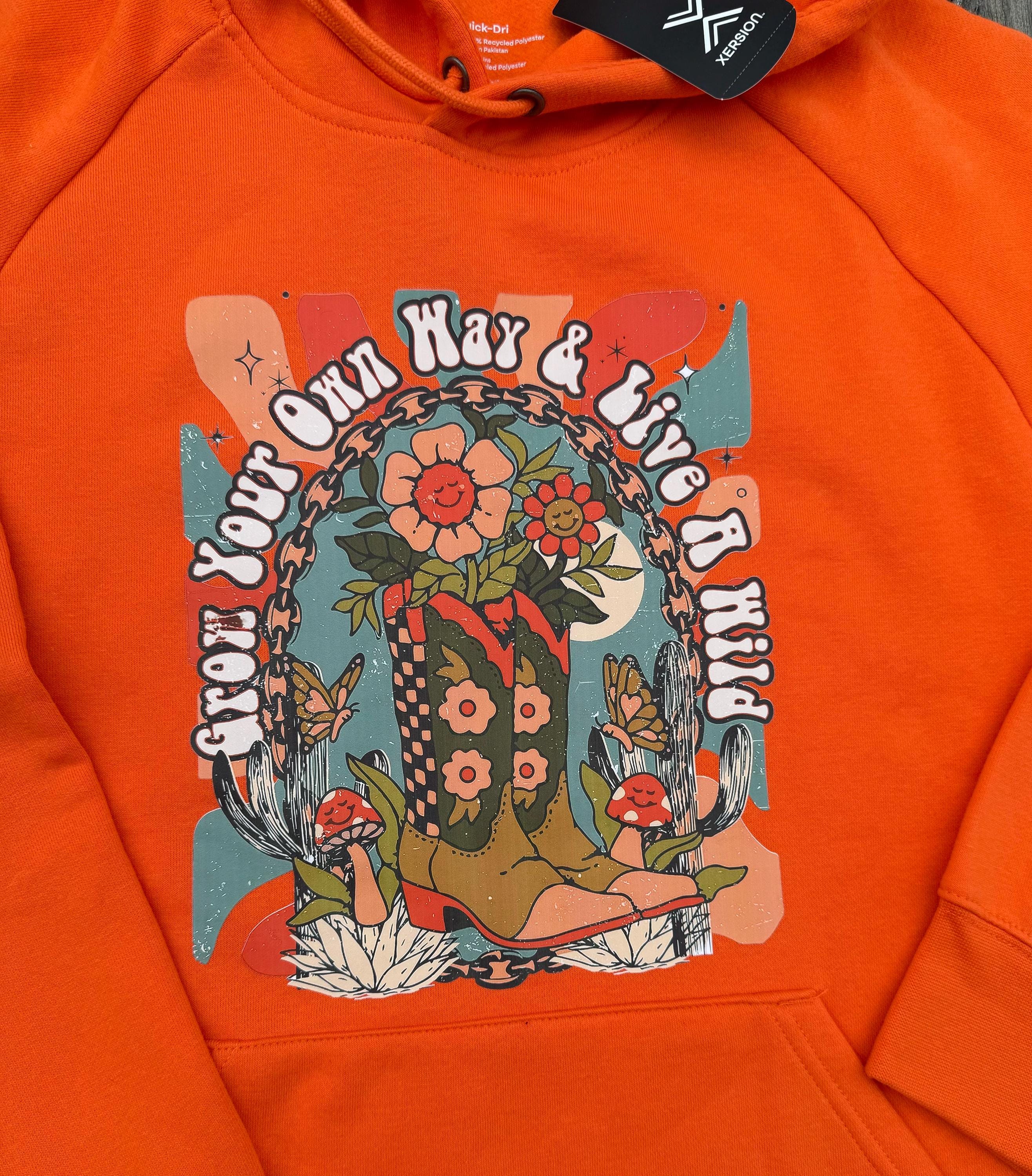 Close-up of an orange hoodie featuring a colorful retro-style graphic print on the chest with flowers, mushrooms, butterflies.