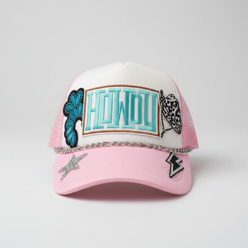 A pink and white trucker hat with 'HOWDY' embroidered in teal on the front, decorated with patches, a silver chain across the brim, and a rhinestone star on the pink brim.