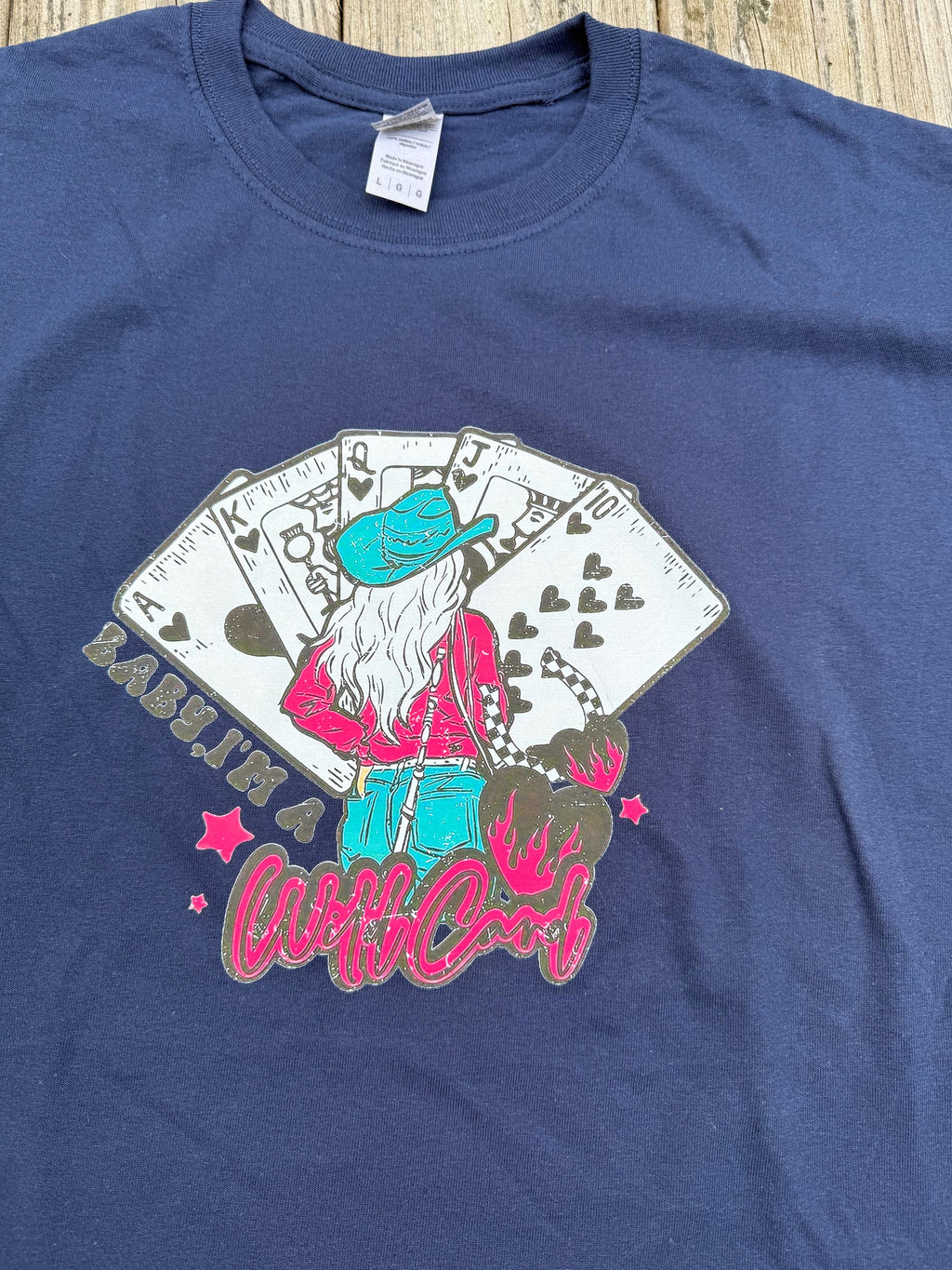 A navy blue short-sleeve t-shirt featuring a colorful graphic print of a woman in a turquoise hat and pink top, seated in front of playing cards, with text in pink script.