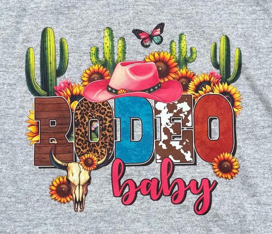 A close-up of a heather gray t-shirt featuring a colorful graphic print with the word 'RODEO' in western-themed patterned letters, a pink cowboy hat, cacti, sunflowers, a butterfly, and a skull, above the text 'baby' in pink script.