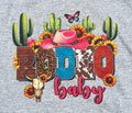 A close-up of a heather gray t-shirt featuring a colorful graphic print with the word 'RODEO' in western-themed patterned letters, a pink cowboy hat, cacti, sunflowers, a butterfly, and a skull, above the text 'baby' in pink script.