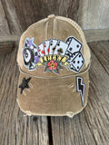 A distressed tan trucker cap with a 'Lucky' playing card theme, featuring playing card patches, dice, a star, and a lightning bolt on a rustic wooden background.