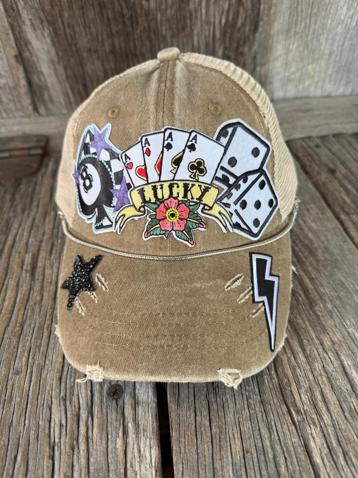 A distressed tan trucker cap with a 'Lucky' playing card theme, featuring playing card patches, dice, a star, and a lightning bolt on a rustic wooden background.