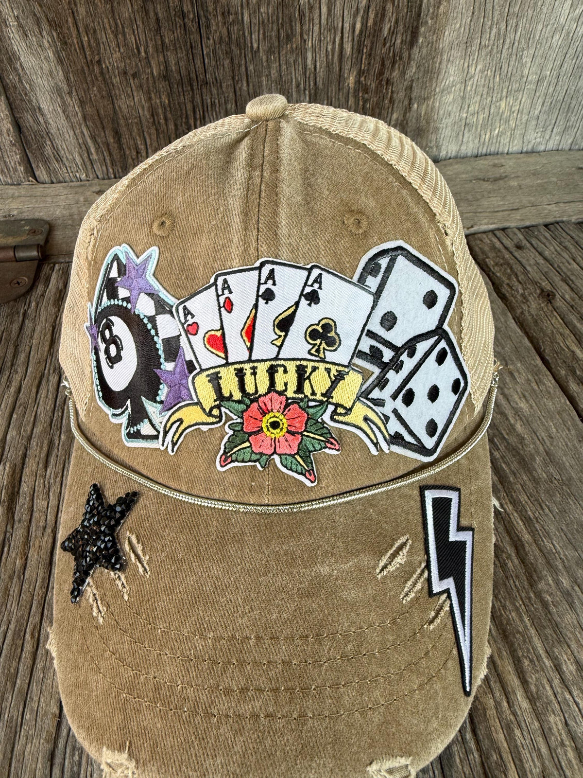 A distressed tan trucker cap with a 'Lucky' poker theme patch on the front, featuring playing cards, dice, a 8-ball, a flower, and embroidered stars and lightning bolts on the brim, displayed on a rustic wooden surface.