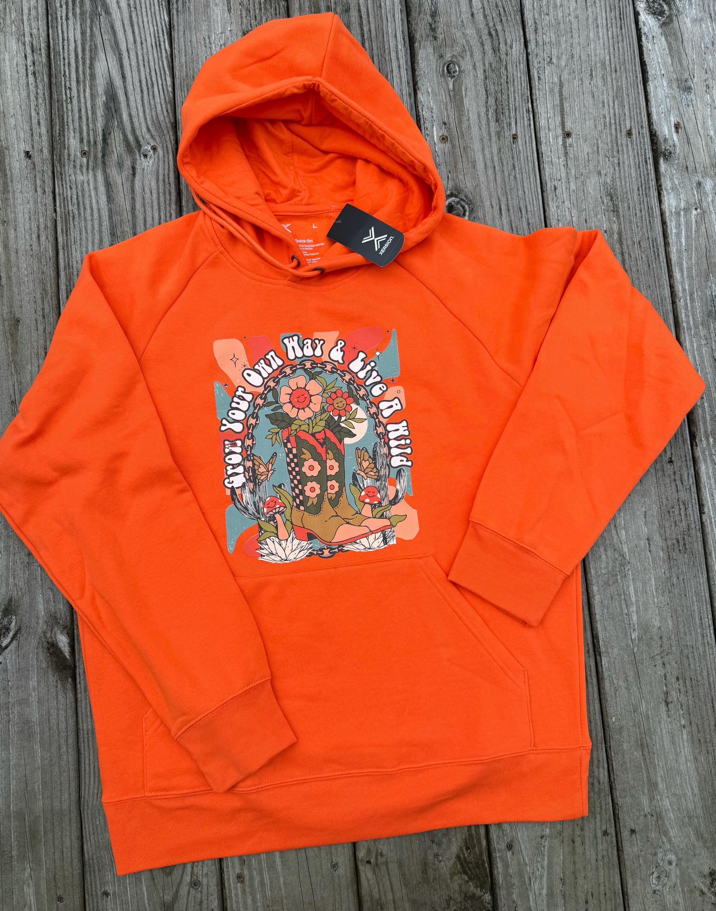 Bright orange pullover hoodie laid flat on a weathered gray wooden plank surface, featuring a large colorful front graphic with floral artwork and stylized text.
