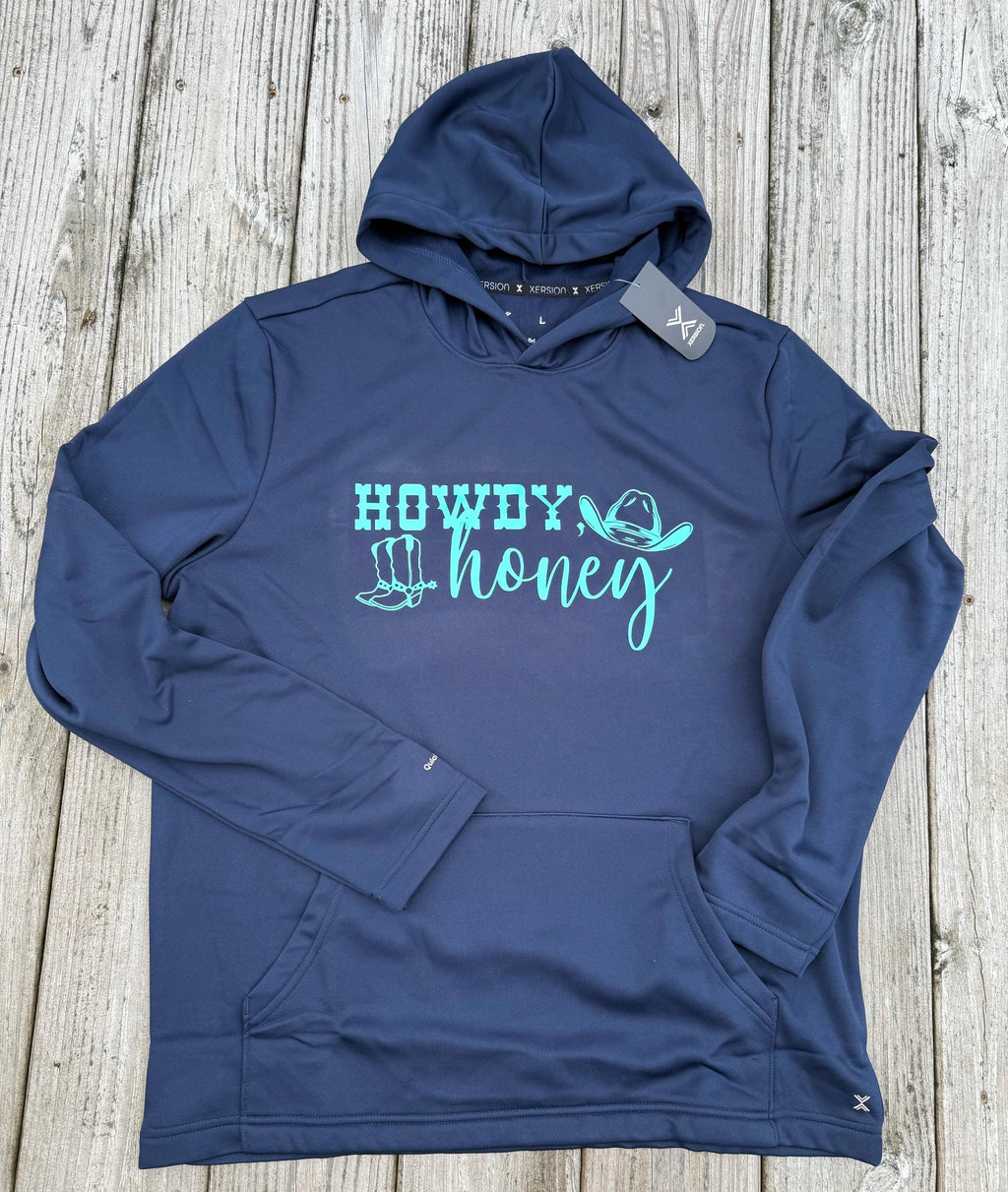 A navy blue athletic hoodie with a turquoise graphic reading 'HOWDY honey' and small hat illustrations, laid flat on a light wood plank background. A gray hangtag is attached near the hood.