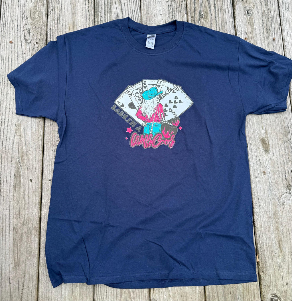 A navy blue short-sleeve graphic t-shirt laid flat on a rustic wooden plank surface. The graphic features a stylized cartoon character with a turquoise hat and pink clothing playing cards, with the word 'Wildcard' written in pink script below.