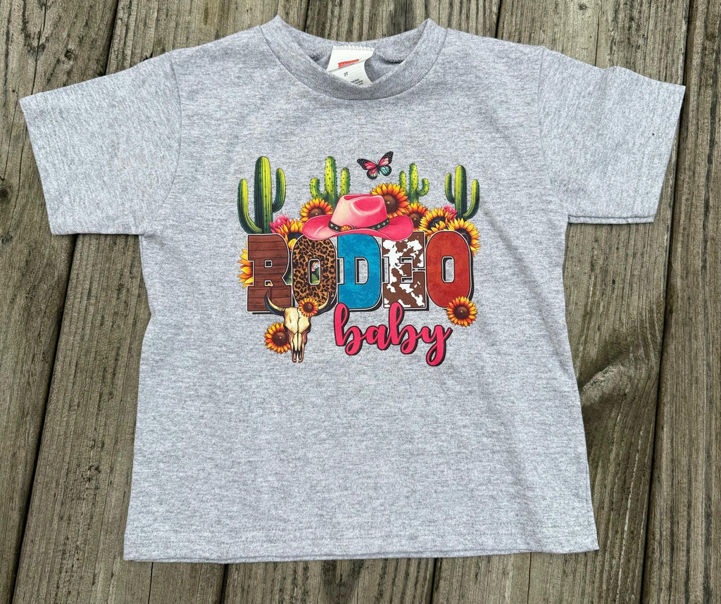 A heather gray toddler t-shirt with a colorful 'Rodeo Baby' graphic print, laid flat on a rustic wooden plank background.