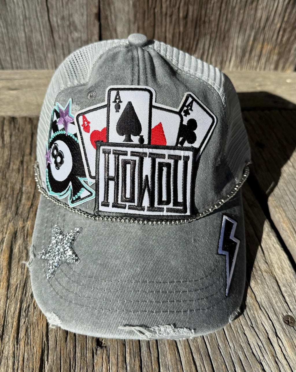 A distressed gray trucker cap with mesh back panels, featuring a large front patch with playing card suits and the word 'HOWDY', plus a 8-ball patch, star embellishment, and lightning bolt patch on the brim, displayed on a rustic wooden surface.