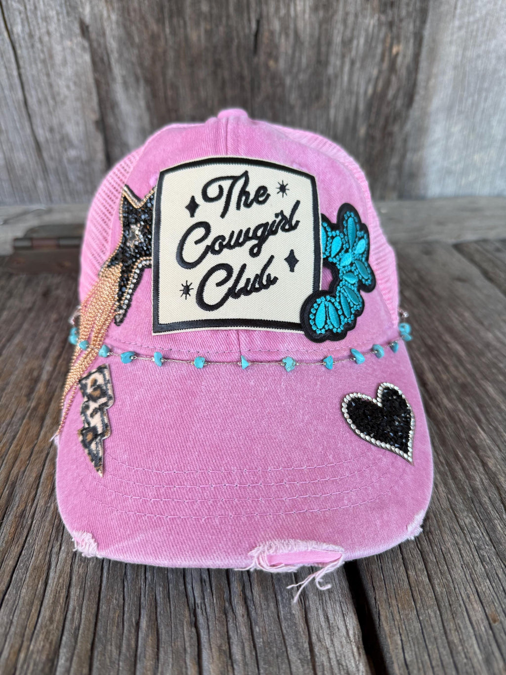 A pink distressed trucker-style cap with a white rectangular patch reading 'The Cowgirl Club' and black script, decorated with a turquoise sequined cowboy hat patch, a black heart patch, and small turquoise beads along the front band, photographed on a rustic wooden surface.