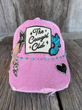 A pink distressed trucker-style cap with a white rectangular patch reading 'The Cowgirl Club' and black script, decorated with a turquoise sequined cowboy hat patch, a black heart patch, and small turquoise beads along the front band, photographed on a rustic wooden surface.