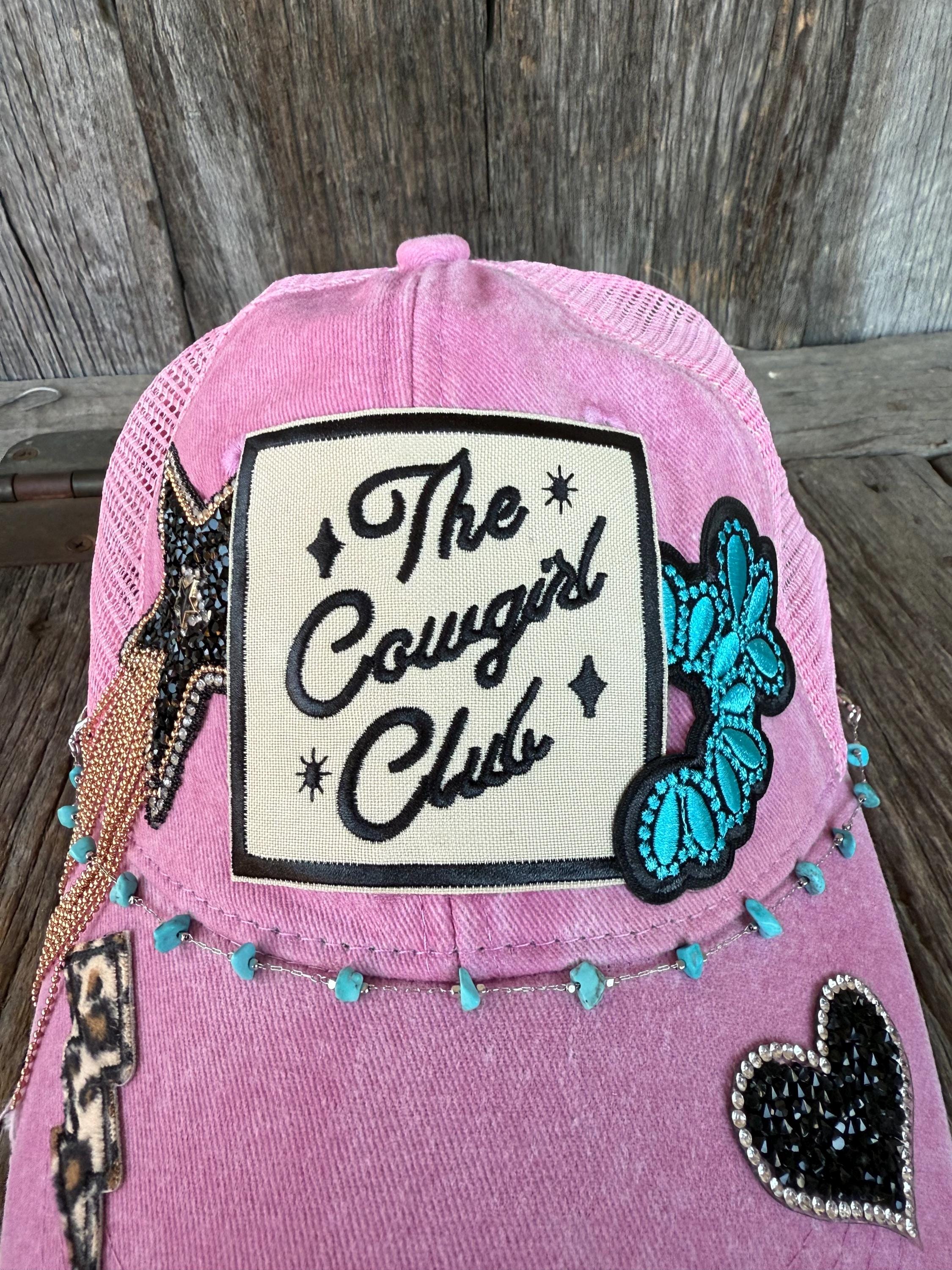 A pink trucker-style cap with a cream patch reading 'The Cowgirl Club' and multiple western-themed embellishments, including a turquoise butterfly patch, leopard-print star, and black heart patch, displayed against a rustic wooden background.