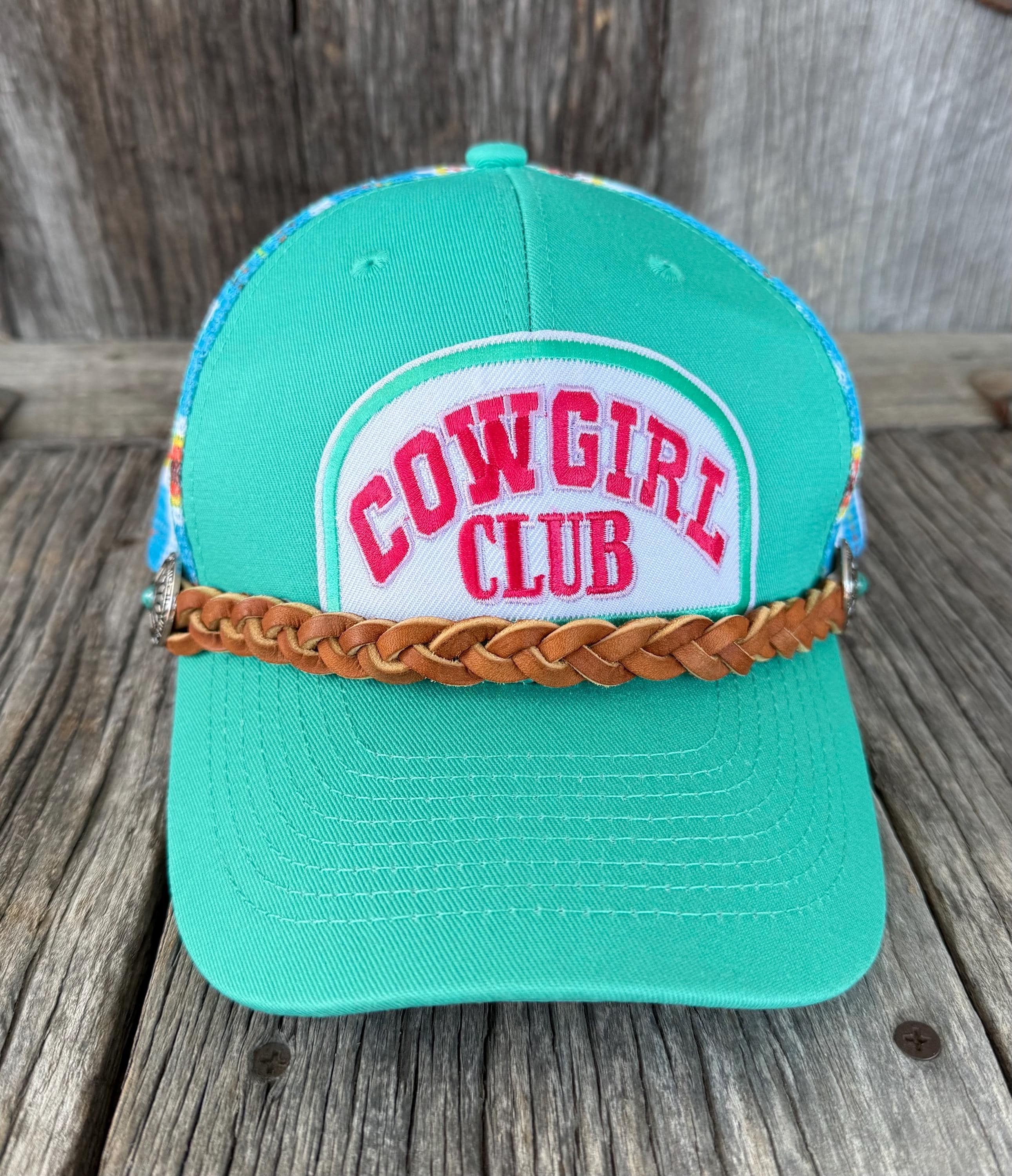 A turquoise trucker-style cap with a white front patch reading 'COWGIRL CLUB' in red letters, a tan braided headband across the brim, and multicolor patterned side panels, photographed on a rustic wooden surface.