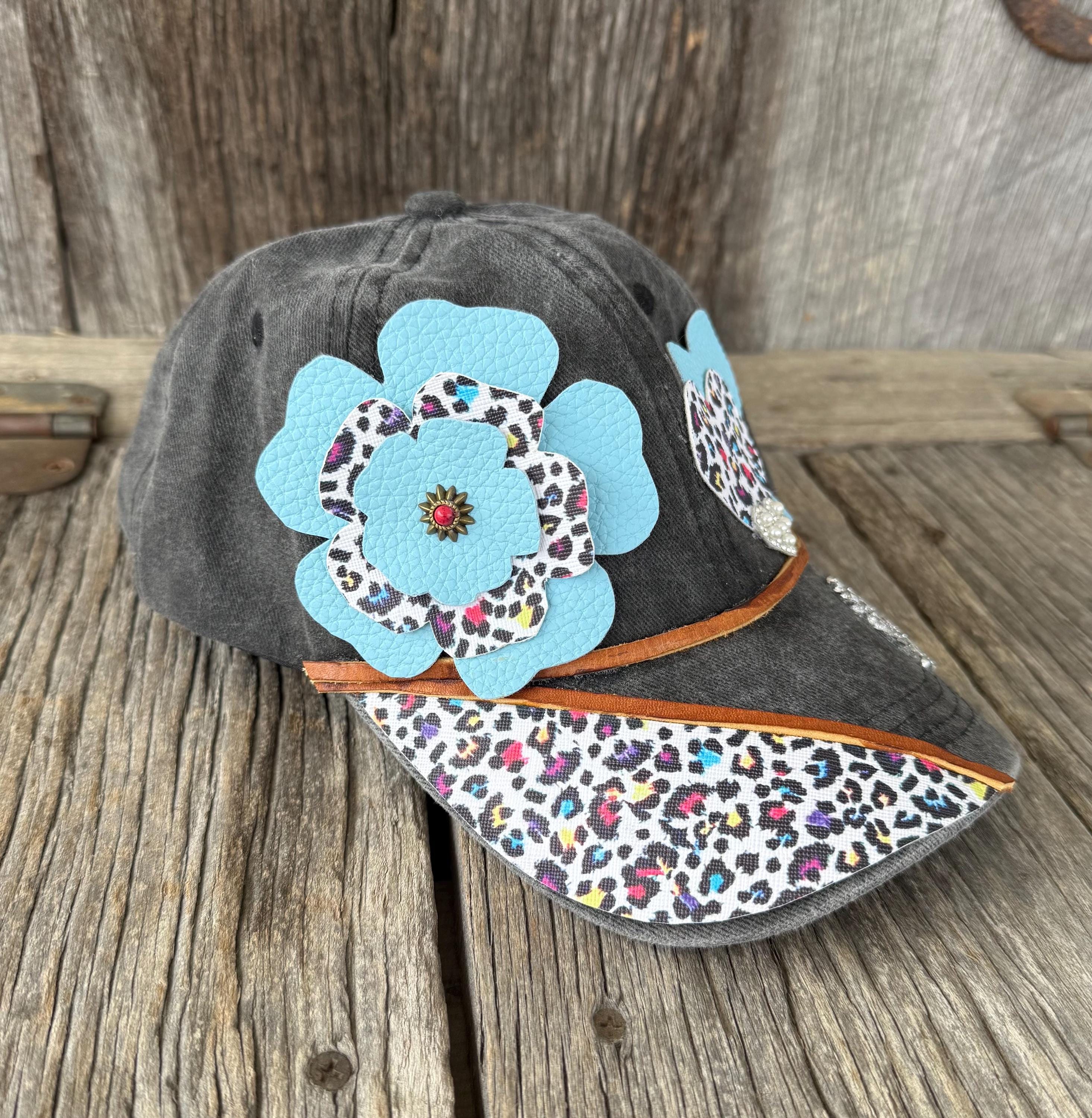 A dark gray baseball cap with a large light blue flower applique and a multicolor leopard print brim, displayed on a rustic wooden surface.