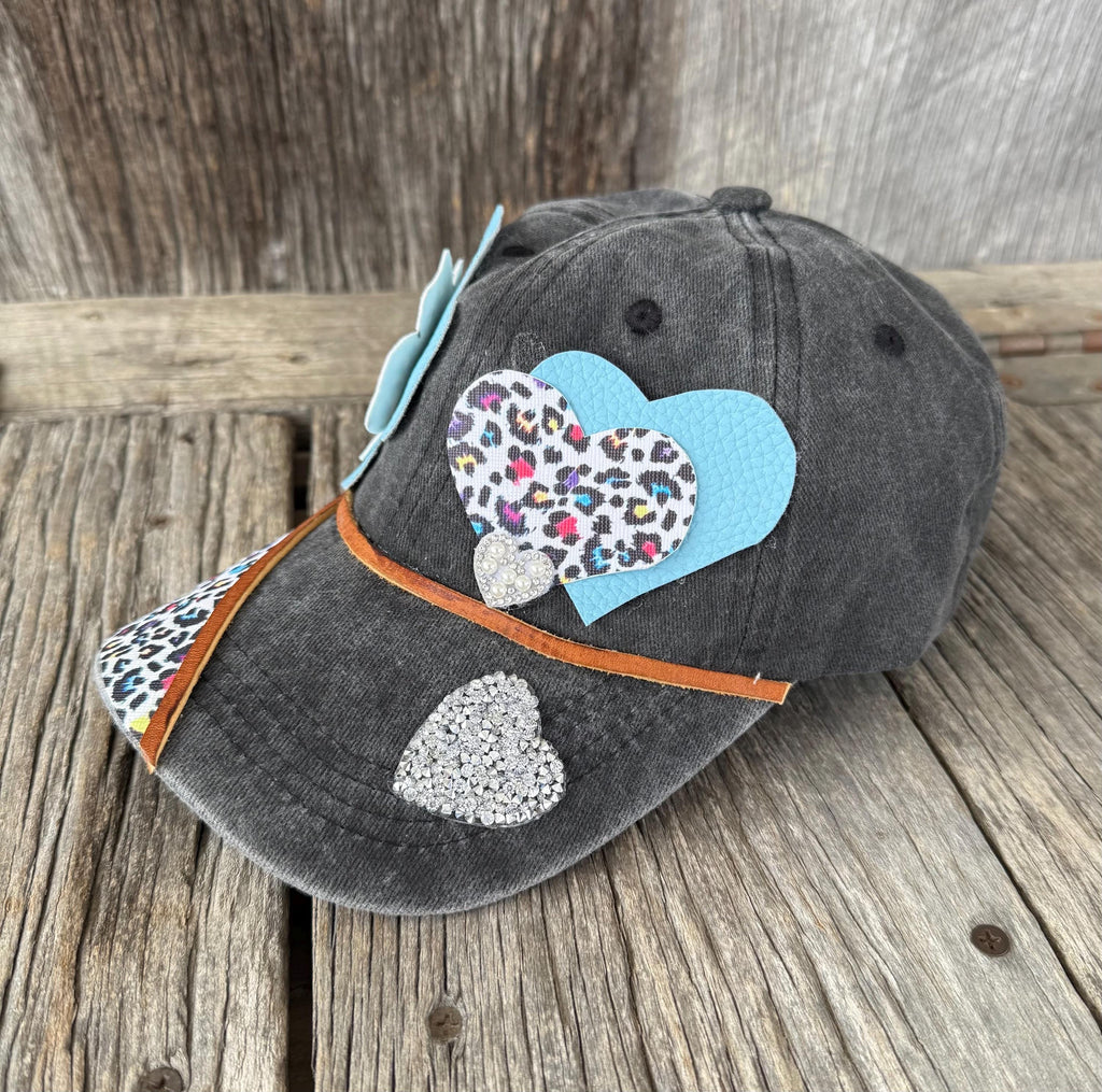 A dark gray washed baseball cap with a leopard-print brim, decorated with a light blue bow, a large light blue and leopard-print heart patch, a small silver heart patch, and an orange cord, displayed on a rustic wooden surface.
