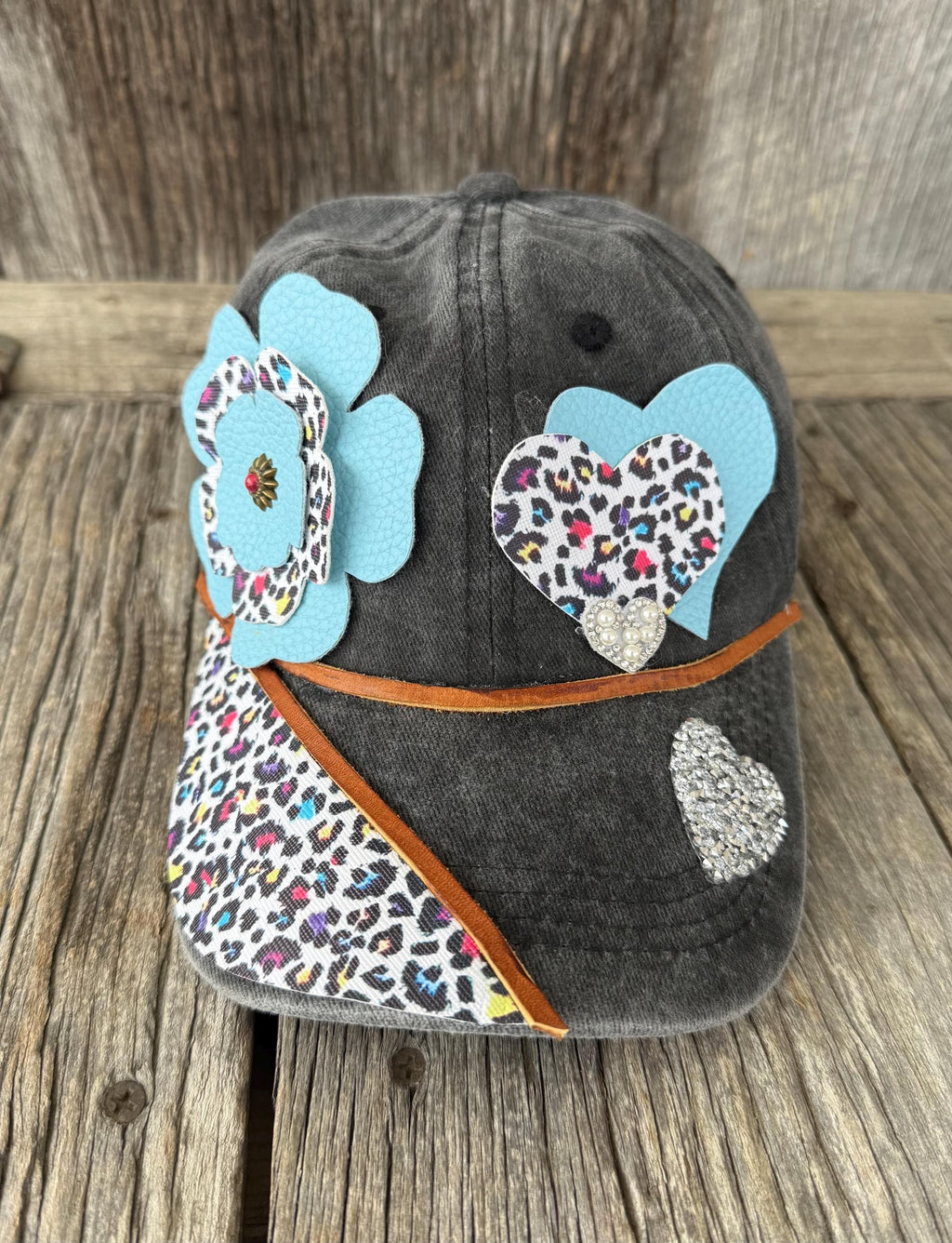A dark gray washed baseball cap decorated with light blue felt flowers, leopard-print heart patches, a leopard-print visor panel, and a silver rhinestone heart, displayed on a rustic wooden surface.