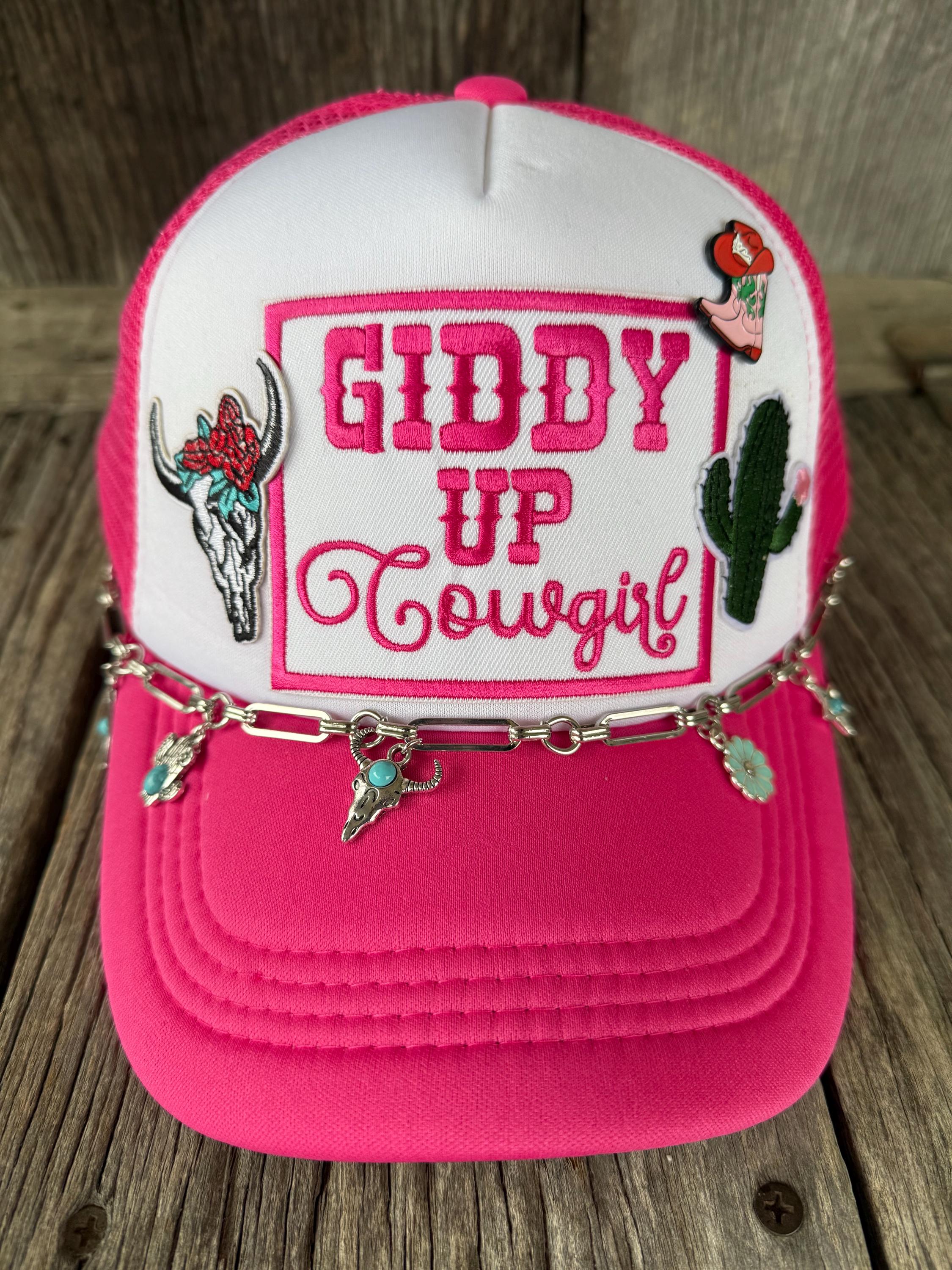 A pink and white trucker hat with 'GIDDY UP Cowgirl' embroidered on the front, featuring a silver chain with turquoise charms draped across the brim, set against a rustic wooden background.