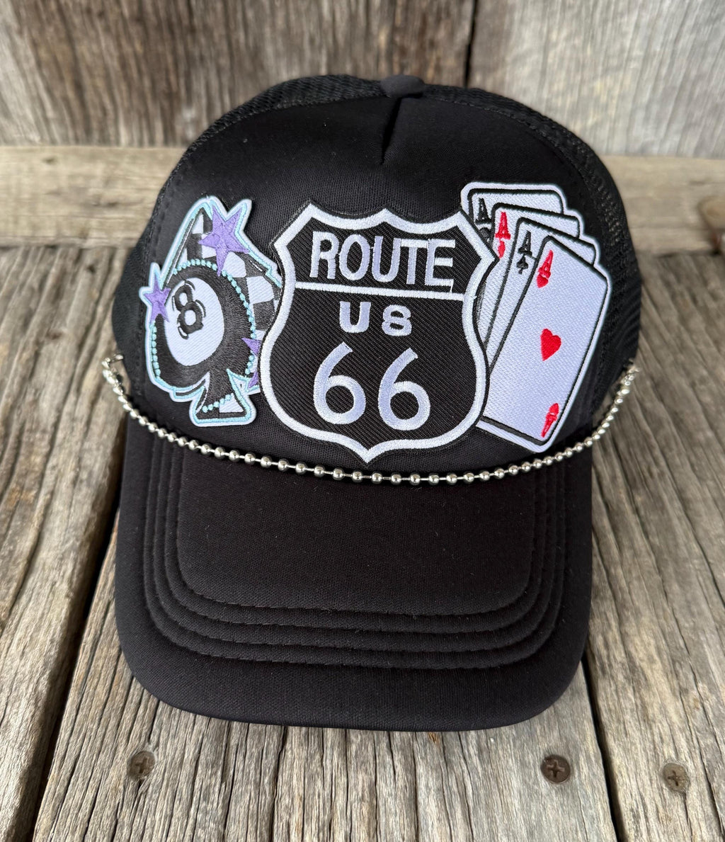 A black trucker-style cap with embroidered patches including a Route 66 shield, an 8-ball with stars, and playing cards, resting on a rustic wooden surface.