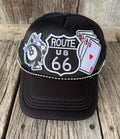 A black trucker-style cap with embroidered patches including a Route 66 shield, an 8-ball with stars, and playing cards, resting on a rustic wooden surface.