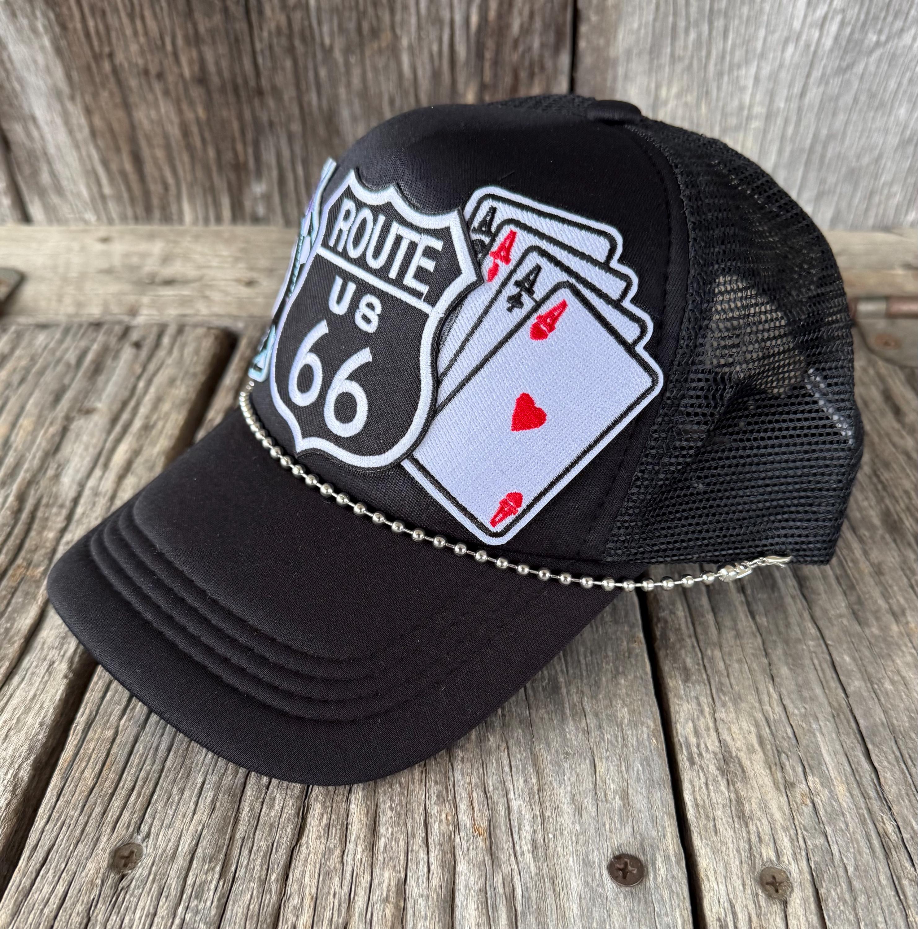 A black trucker-style cap with a curved brim, black mesh back, and an embroidered patch featuring a Route 66 shield and playing cards, resting on a rustic wooden surface.