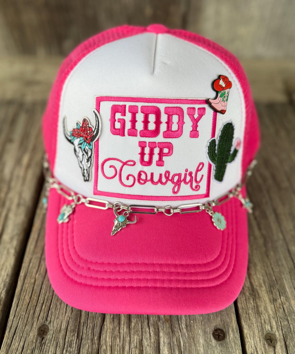 A hot pink and white trucker hat with western-themed embroidery, including a bull skull, cactus, and the text 'GIDDY UP Cowgirl'. A silver chain with turquoise-colored cowgirl hat charms drapes across the front.