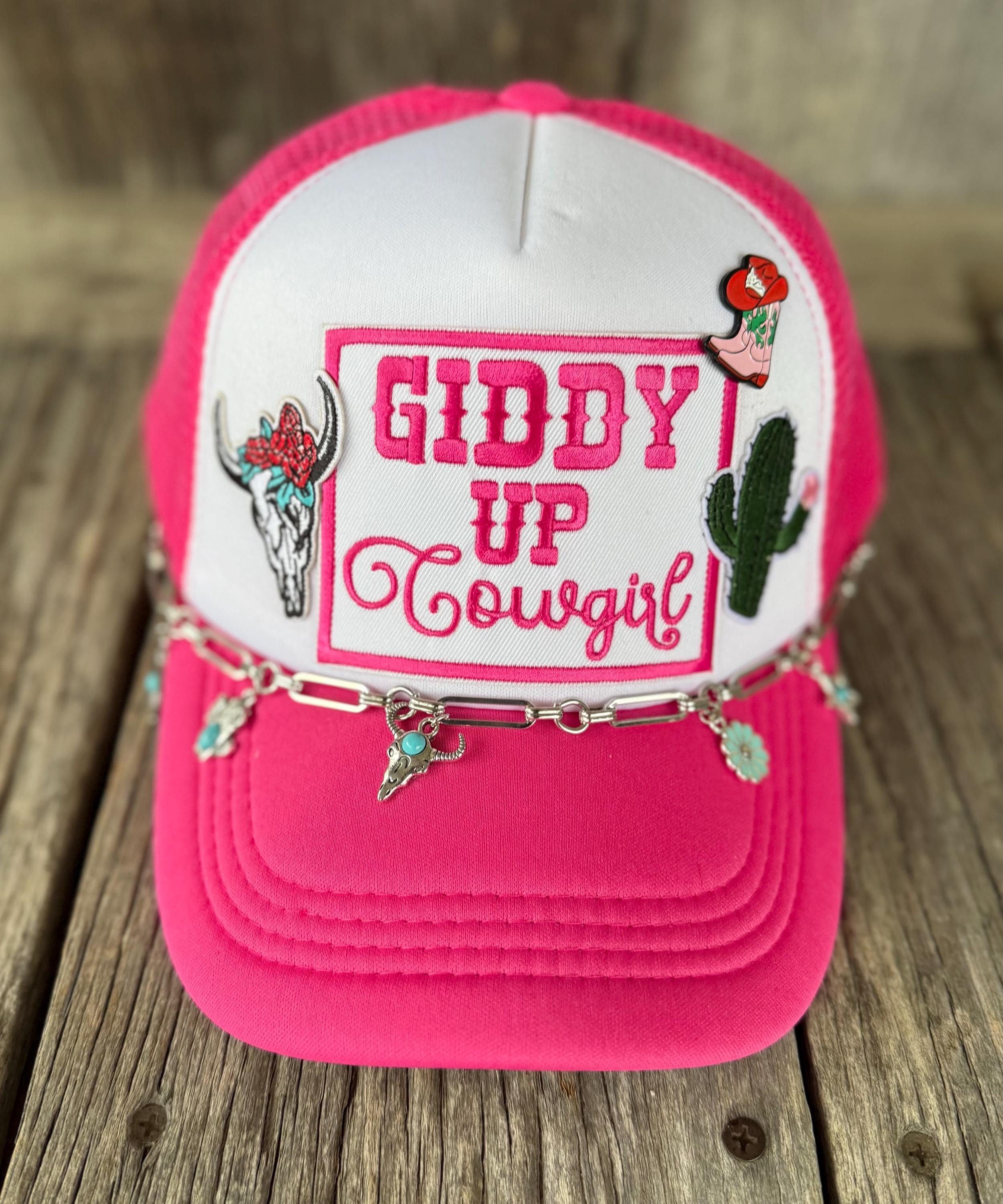 A hot pink and white trucker hat with western-themed embroidery, including a bull skull, cactus, and the text 'GIDDY UP Cowgirl'. A silver chain with turquoise-colored cowgirl hat charms drapes across the front.