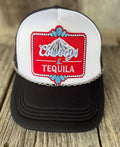 A black and white trucker hat with a red embroidered patch on the front that reads 'Cowboys & Tequila' with a mountain graphic, resting on a rustic wooden surface.