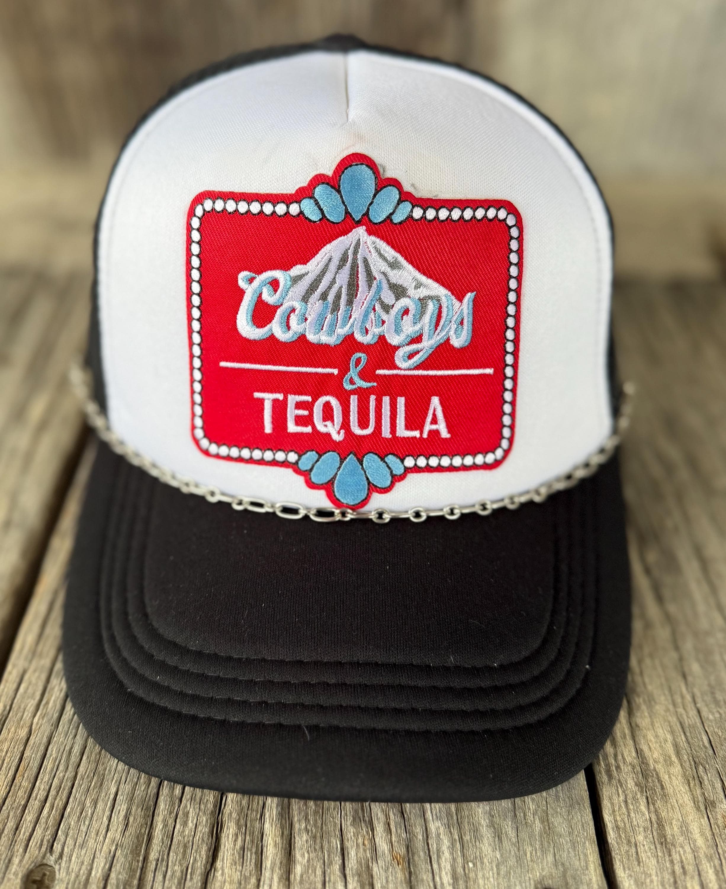 A black and white trucker hat with a red embroidered patch on the front that reads 'Cowboys & Tequila' with a mountain graphic, resting on a rustic wooden surface.