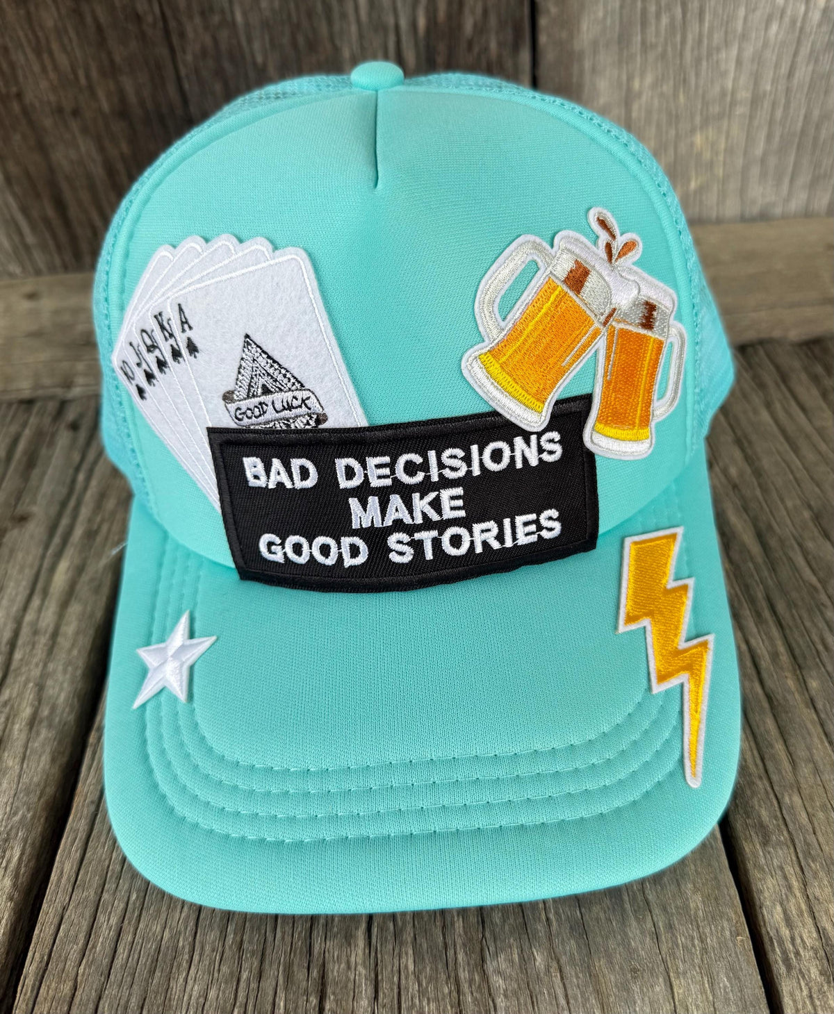 A turquoise trucker cap with various embroidered patches, including a black patch that reads 'BAD DECISIONS MAKE GOOD STORIES', playing cards, clinking beer mugs, a yellow lightning bolt, and a white star, displayed on a rustic wooden background.