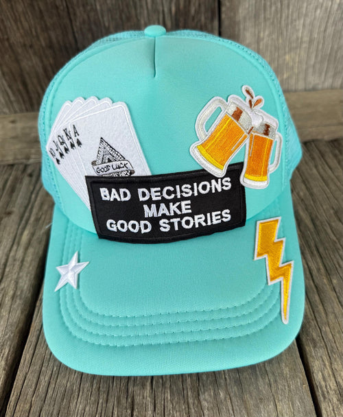 A turquoise trucker cap with various embroidered patches, including a black patch that reads 'BAD DECISIONS MAKE GOOD STORIES', playing cards, clinking beer mugs, a yellow lightning bolt, and a white star, displayed on a rustic wooden background.