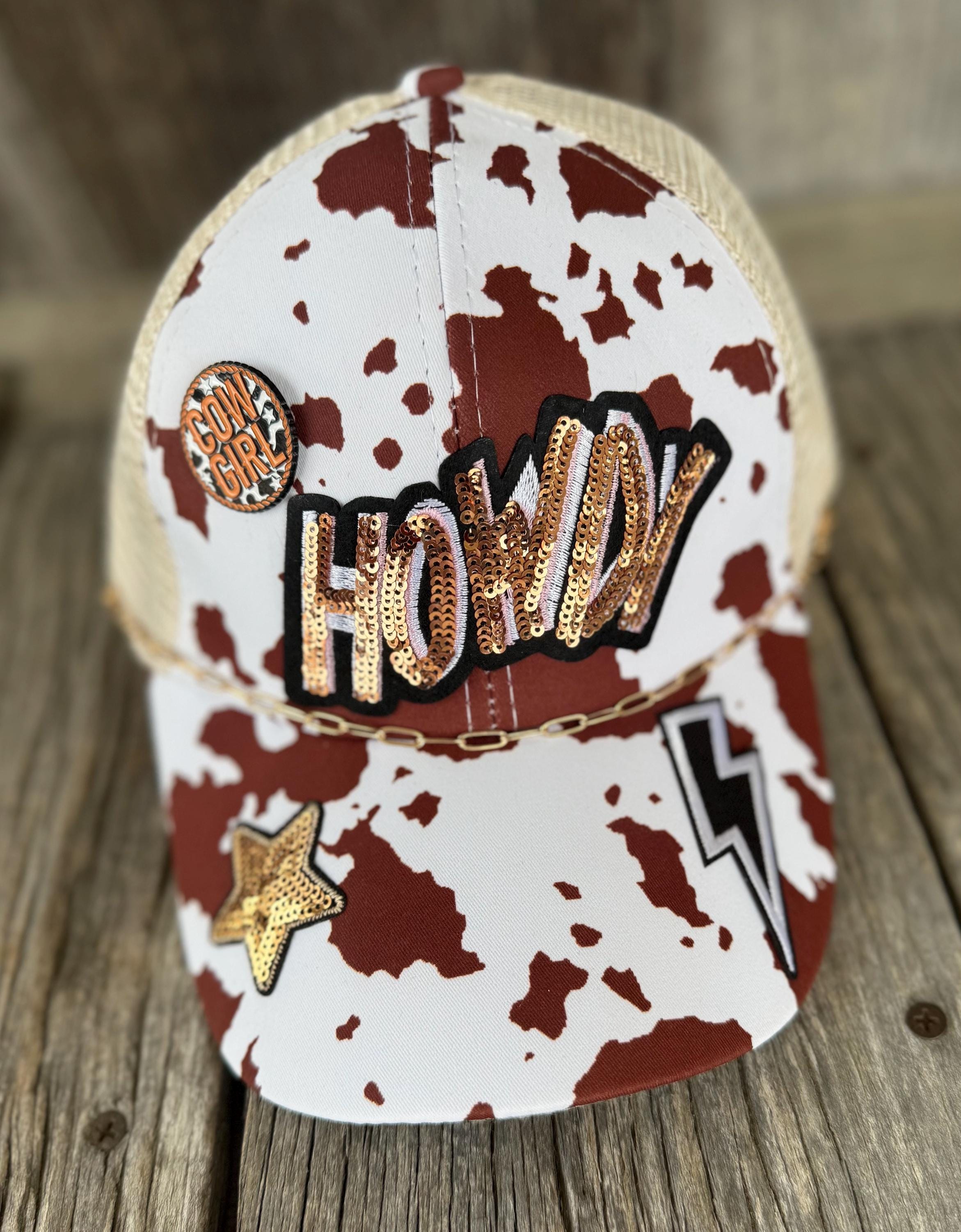 A cow-print trucker hat with a beige mesh back, decorated with sequined 'HOWDY' lettering, a gold star patch, and a lightning bolt patch on a rustic wooden surface.