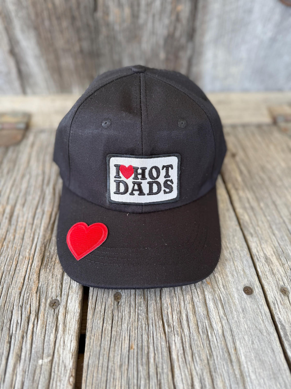 A black baseball cap resting on a rustic wooden surface, featuring a white rectangular patch on the front that reads 'I <heart> HOT DADS' with a red heart, and a red heart patch on the side.