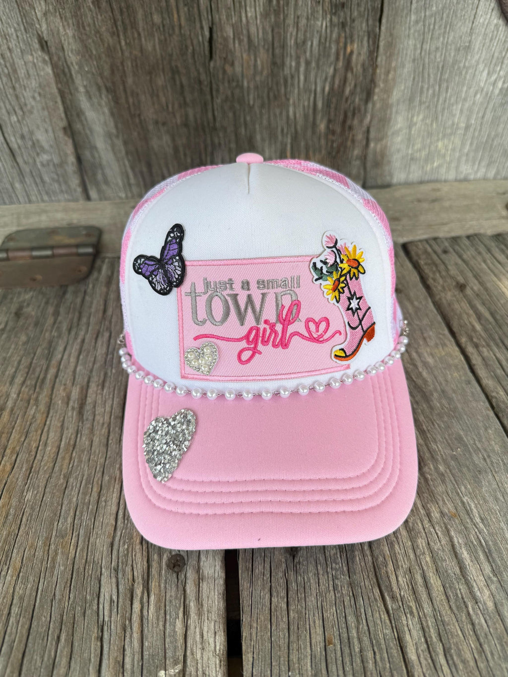 A pink and white trucker hat with western-themed embroidery, rhinestone trim, and decorative patches, displayed on a rustic wooden surface.